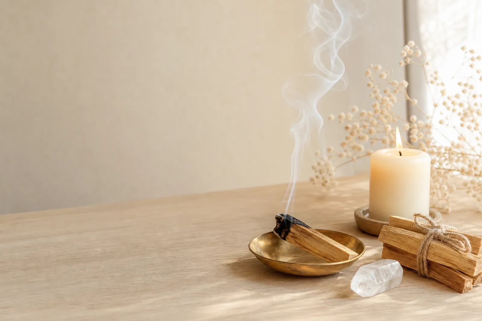 A smouldering palo santo stick in a brass dish with a thin ribbon of smoke, beside dried flowers and a candle